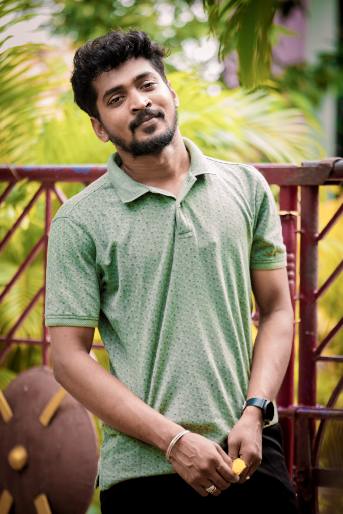 A cheerful young man in a green polo shirt poses casually outdoors against a vibrant garden background.